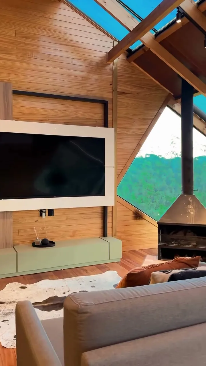 Estar da Cabana Omega com sofá, TV e lareira sob o forro de madeira